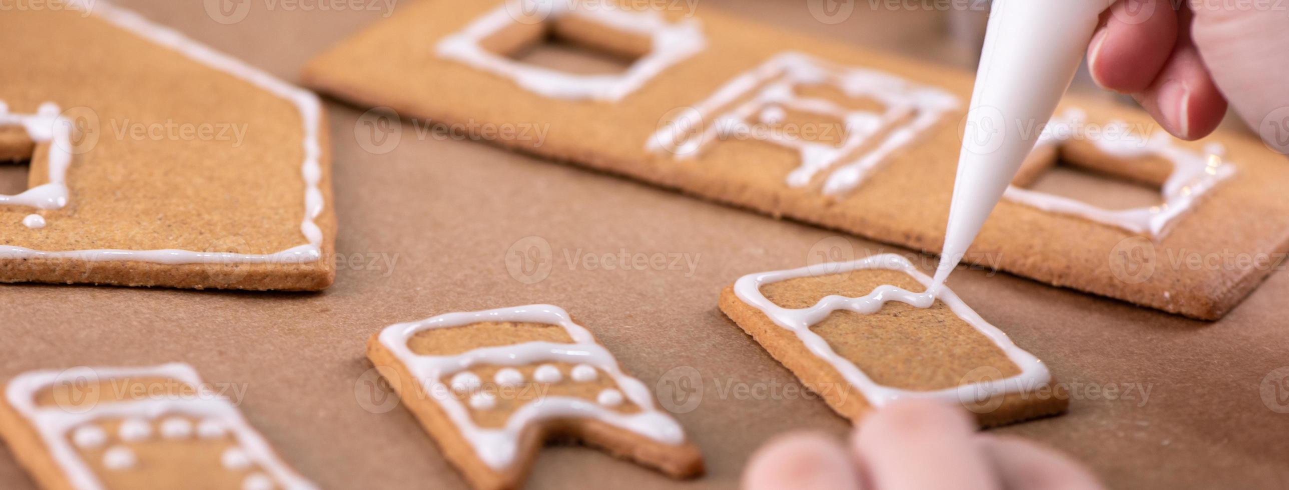 Young woman is decorating Christmas Gingerbread House cookies biscuit at home with frosting topping in icing bag, close up, lifestyle. photo