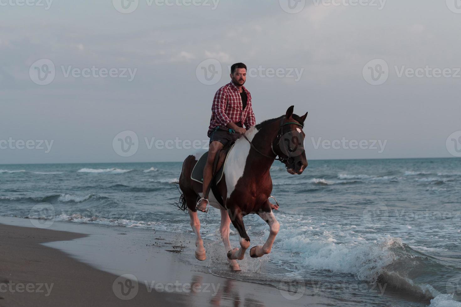 un hombre moderno con ropa de verano disfruta montando a caballo en una hermosa playa de arena al atardecer. enfoque selectivo foto