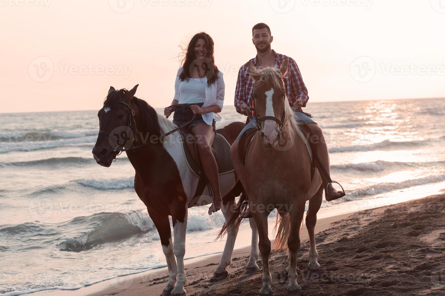 una pareja amorosa vestida de verano montando a caballo en una playa de arena al atardecer. mar y puesta de sol de fondo. enfoque selectivo foto