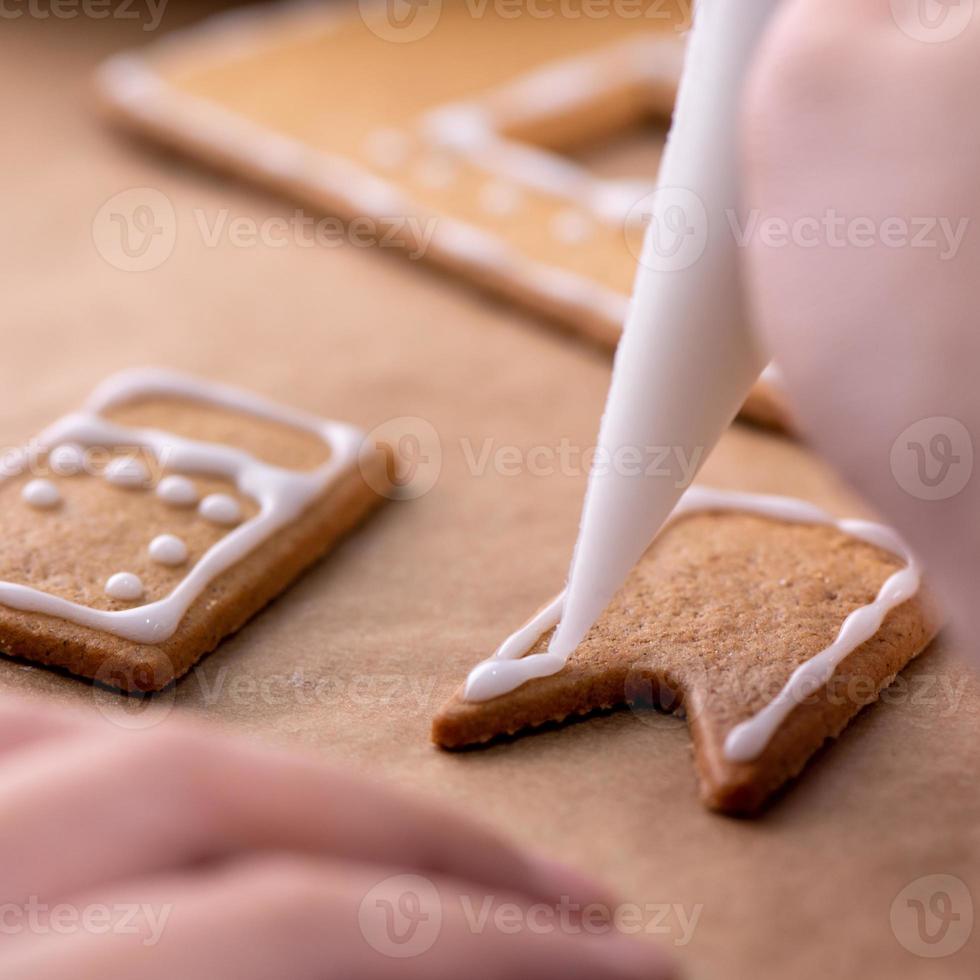 mujer joven está decorando galletas de casa de pan de jengibre de navidad en casa con cobertura de glaseado en bolsa de hielo, primer plano, estilo de vida. foto