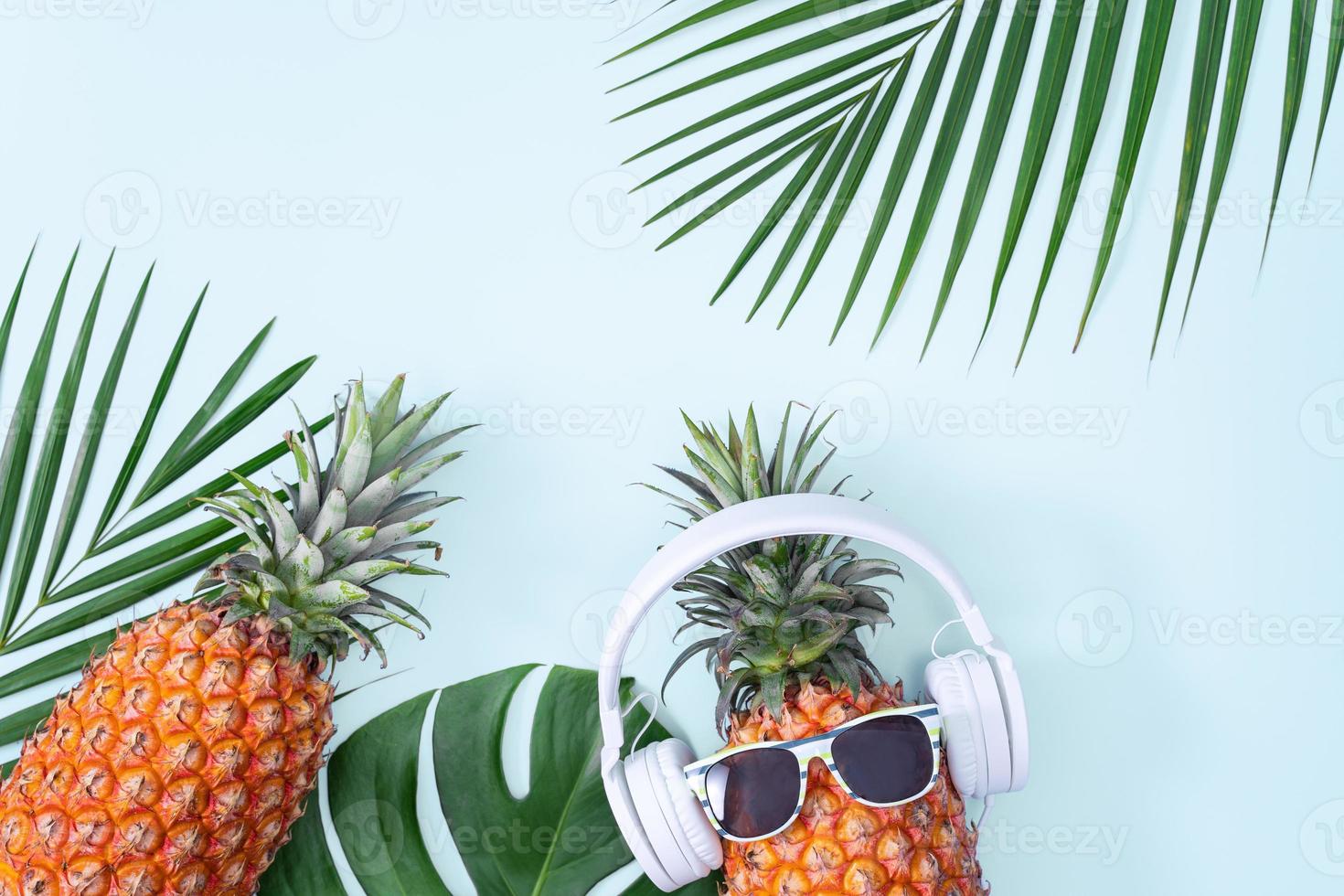 Funny pineapple wearing white headphone, concept of listening music, isolated on blue background with tropical palm leaves, top view, flat lay design. photo