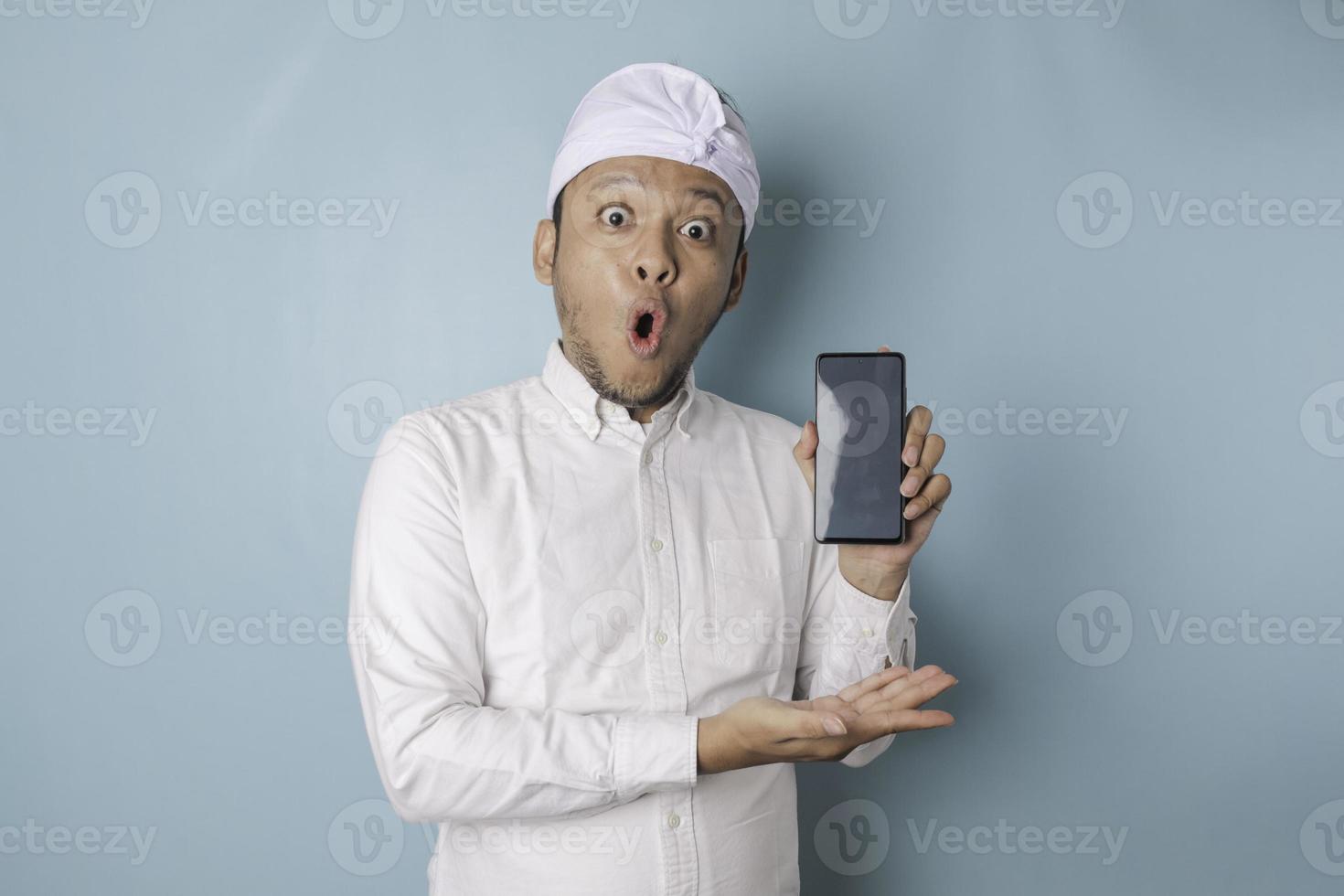 hombre balinés sorprendido con udeng o diadema tradicional y camisa blanca sosteniendo su teléfono inteligente, aislado de fondo azul foto