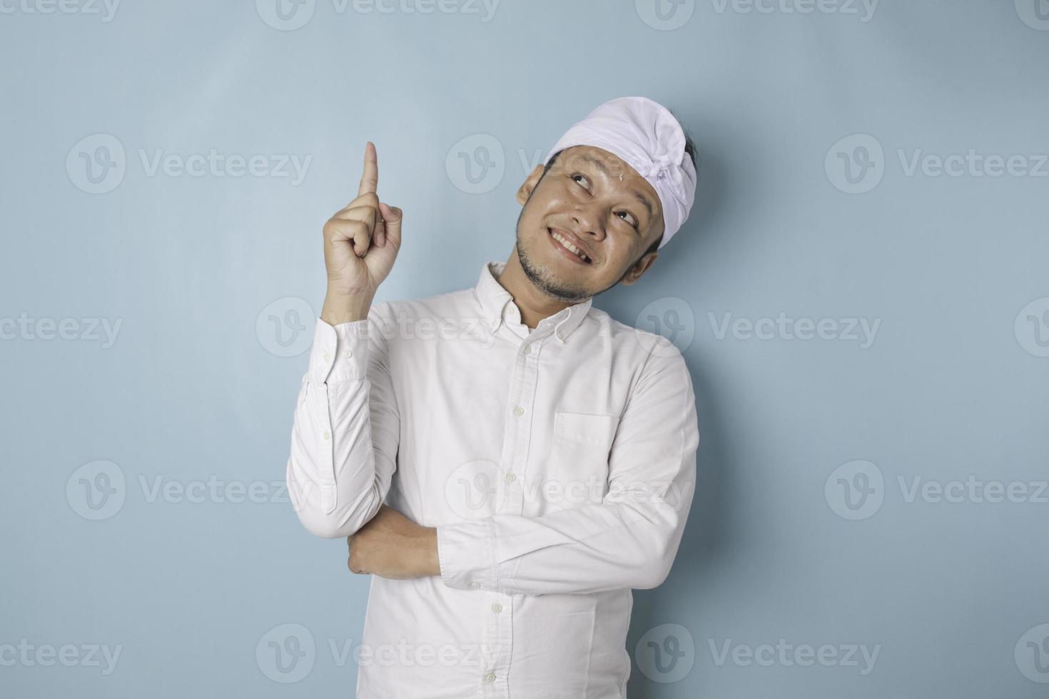 Excited Balinese man wearing udeng or traditional headband and white shirt pointing at the copy space upside him, isolated by blue background photo