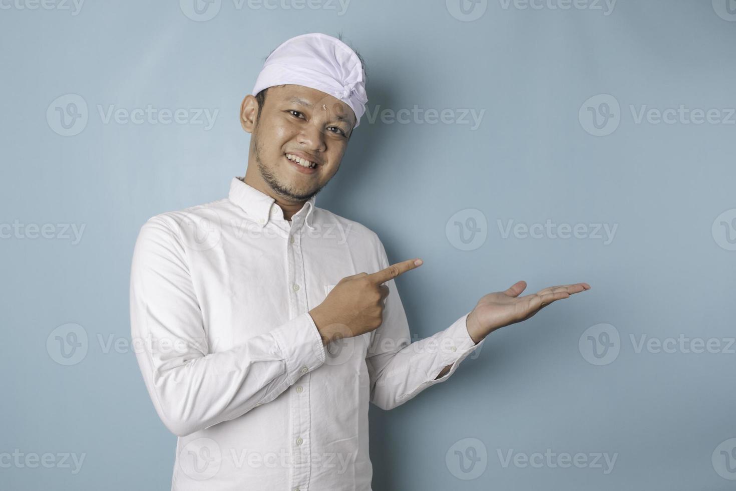 emocionado hombre balinés con udeng o diadema tradicional y pantalones blanco apuntando al espacio de copia a su lado, aislado por fondo azul foto