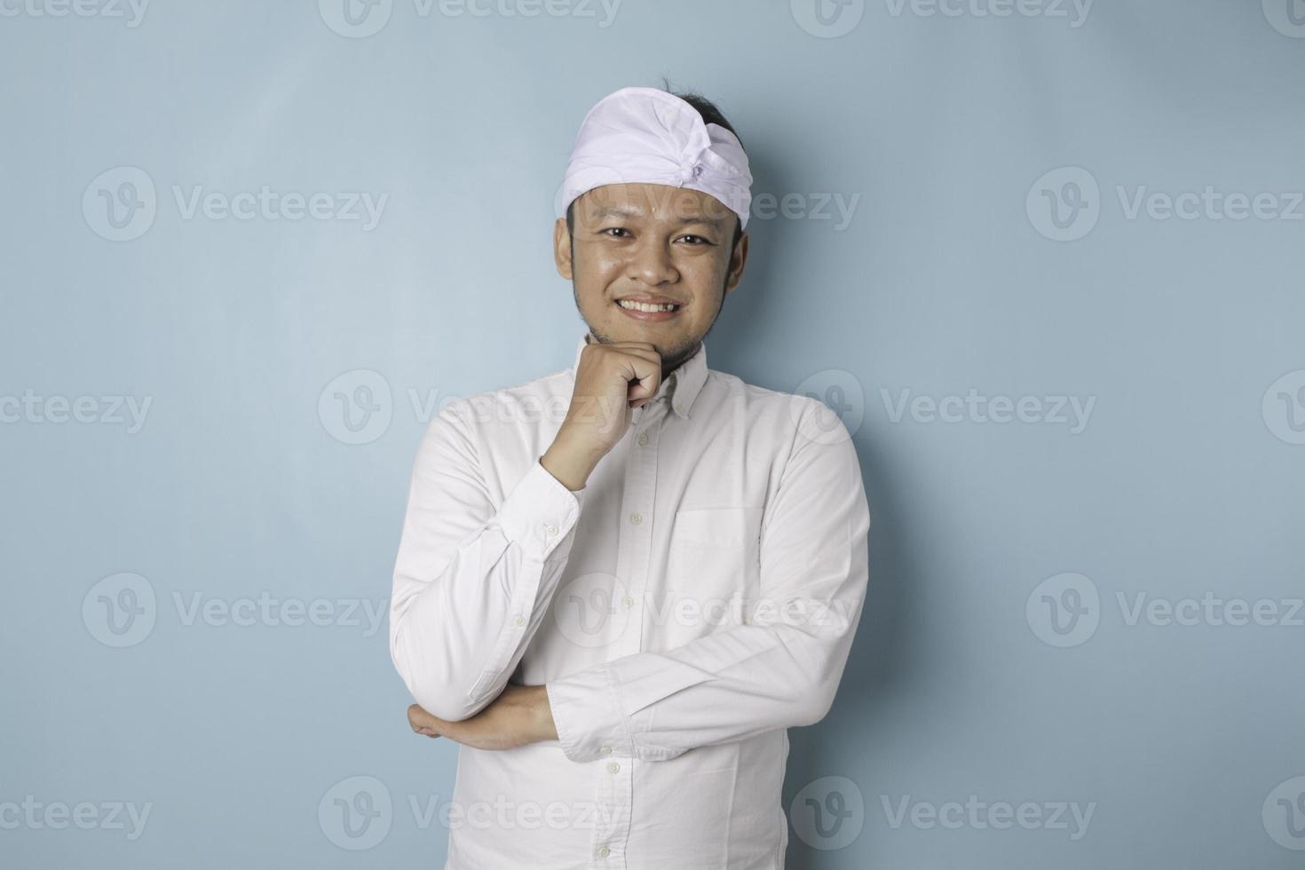 Portrait of a thoughtful young Balinese man wearing udeng or traditional headband and white shirt looking aside while his finger on his chin isolated over blue background photo