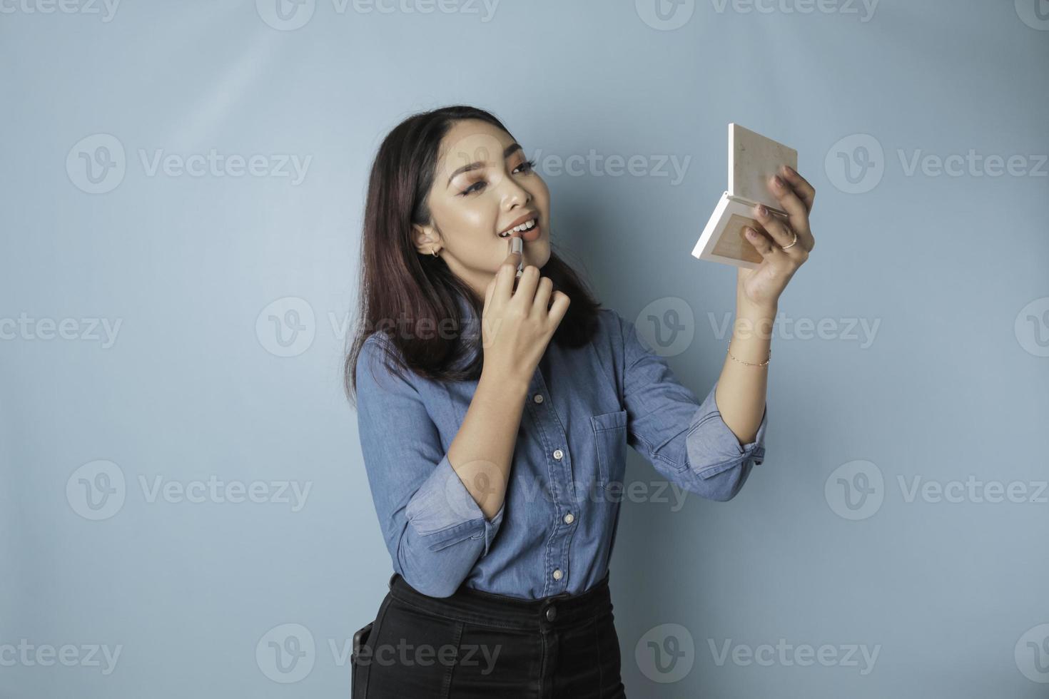 A portrait of a beautiful young Asian woman applying lipstick on her lips, isolated by a blue background photo