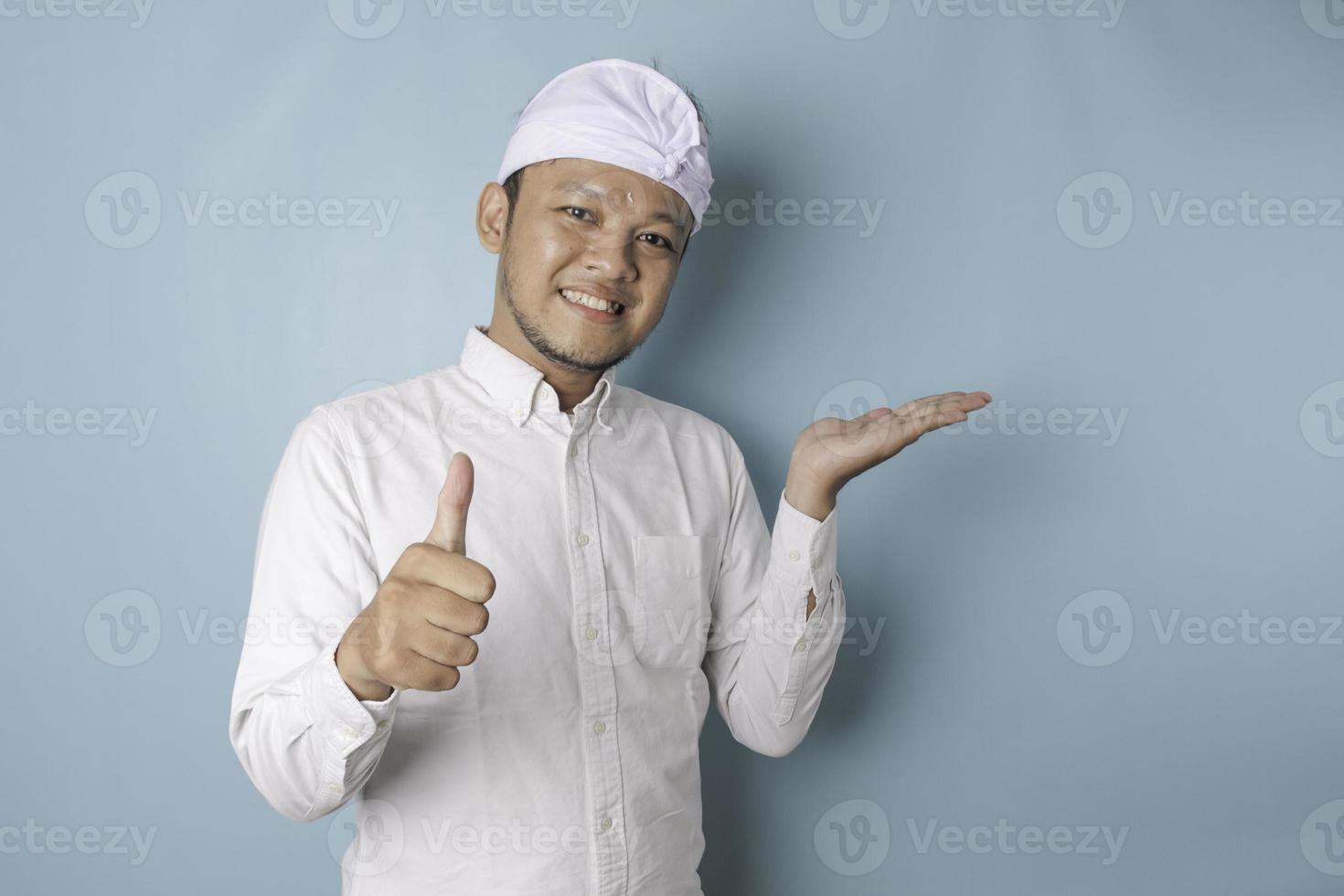 Excited Balinese man wearing udeng or traditional headband and white shirt gives thumbs up hand gesture of approval, isolated by blue background photo