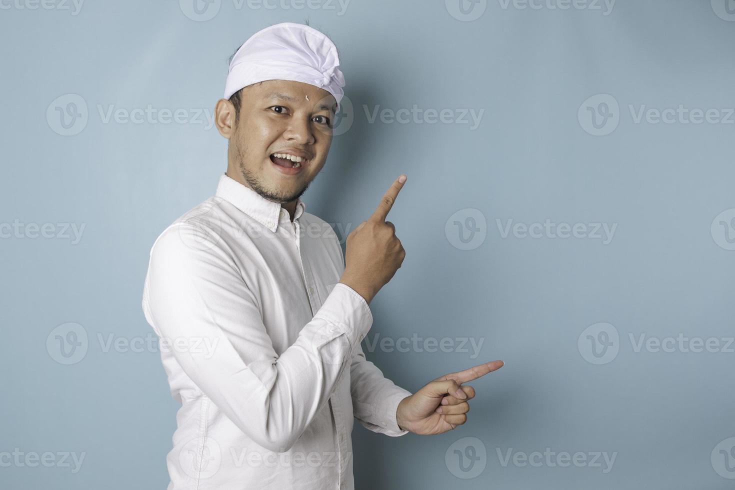 Excited Balinese man wearing udeng or traditional headband and white shirt pointing at the copy space beside him, isolated by blue background photo
