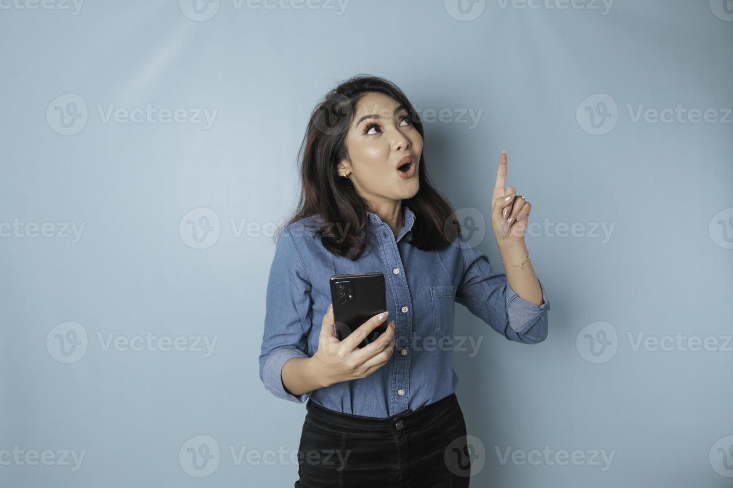 Surprised Asian woman wearing blue shirt pointing at the copy space upside her while holding a smartphone, isolated by blue background photo