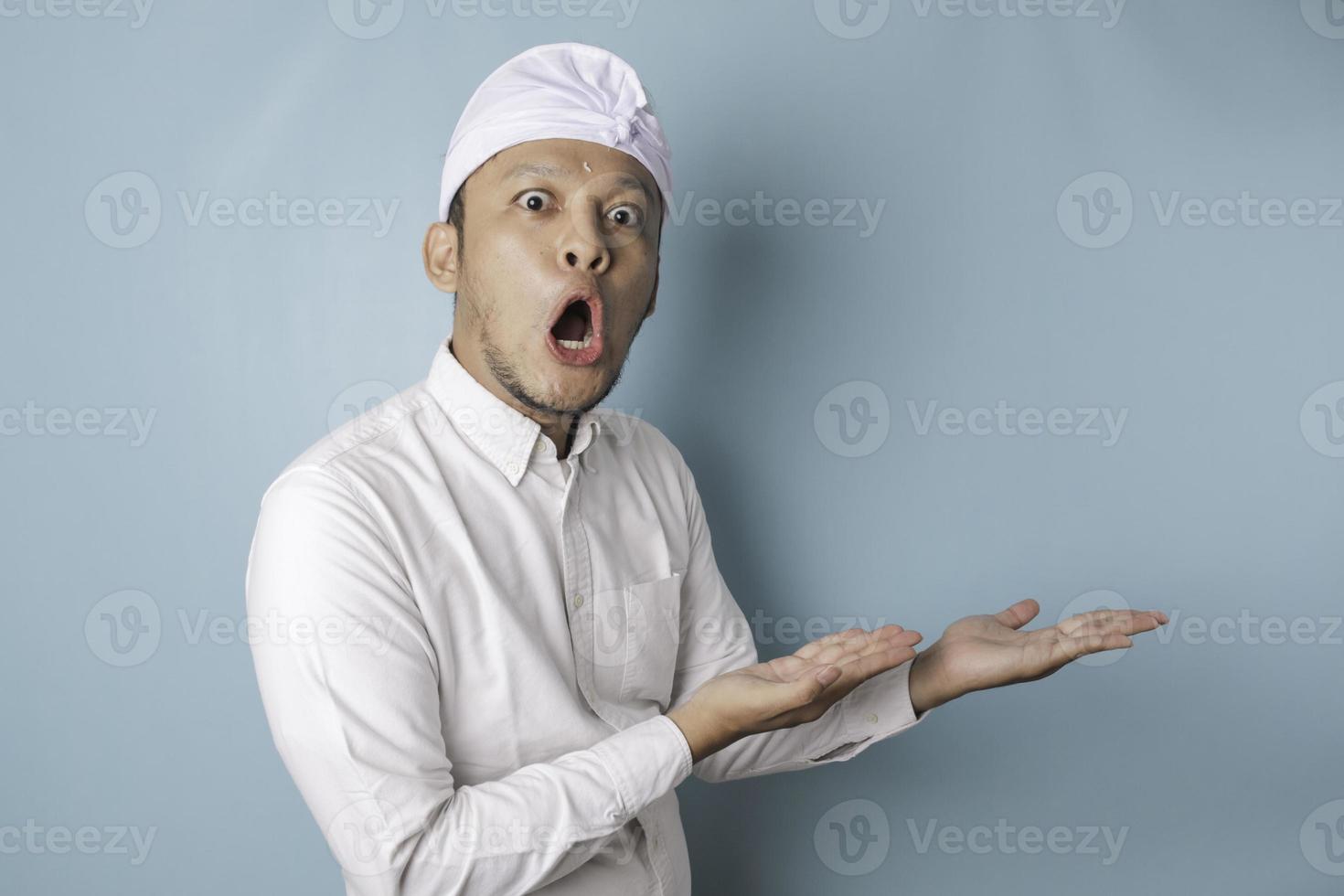 Shocked Balinese man wearing udeng or traditional headband and white shirt pointing at the copy space beside him, isolated by blue background photo