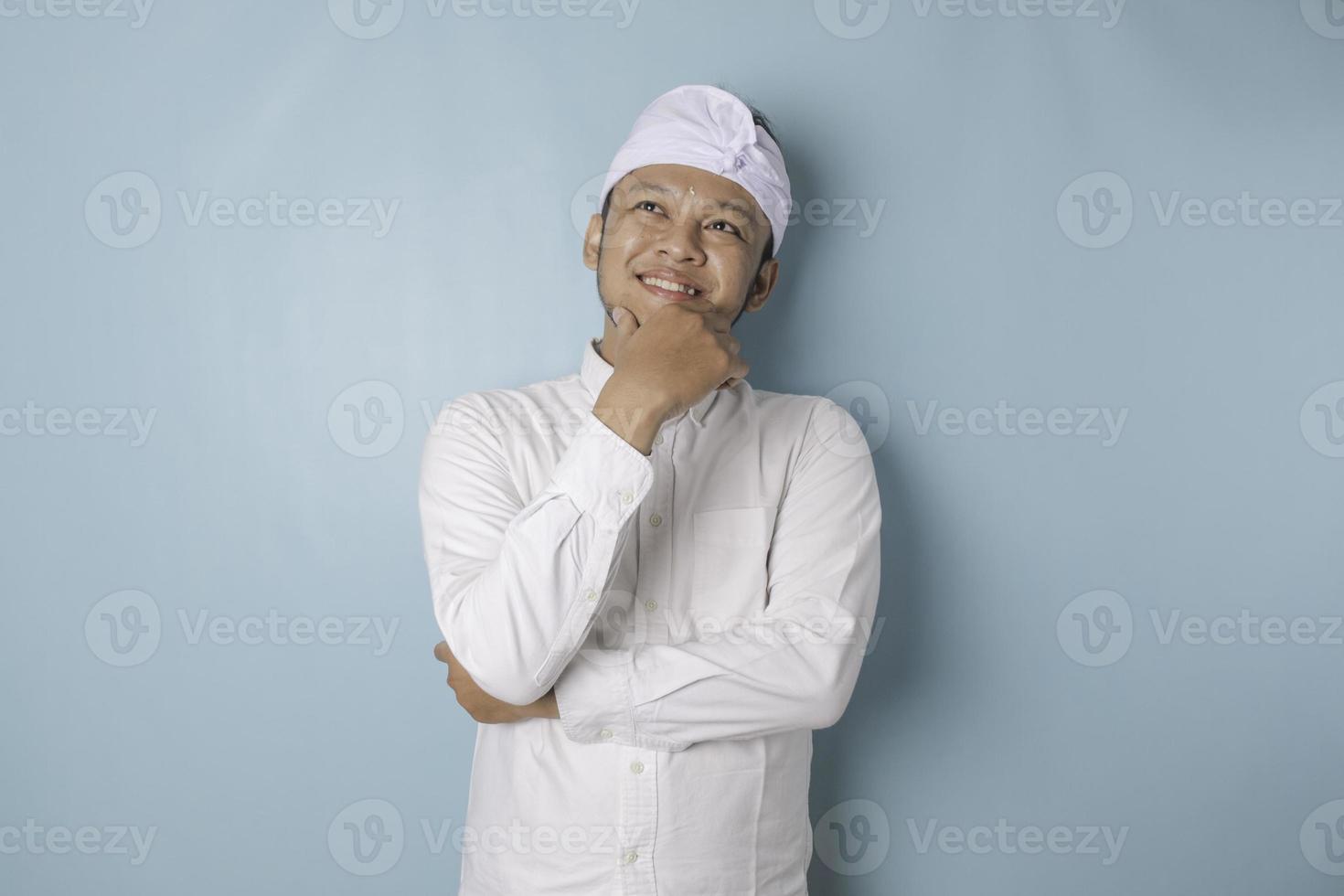 retrato de un joven balinés pensativo que usa udeng o diadema tradicional y camisa blanca mirando a un lado mientras su dedo en la barbilla está aislado sobre fondo azul foto