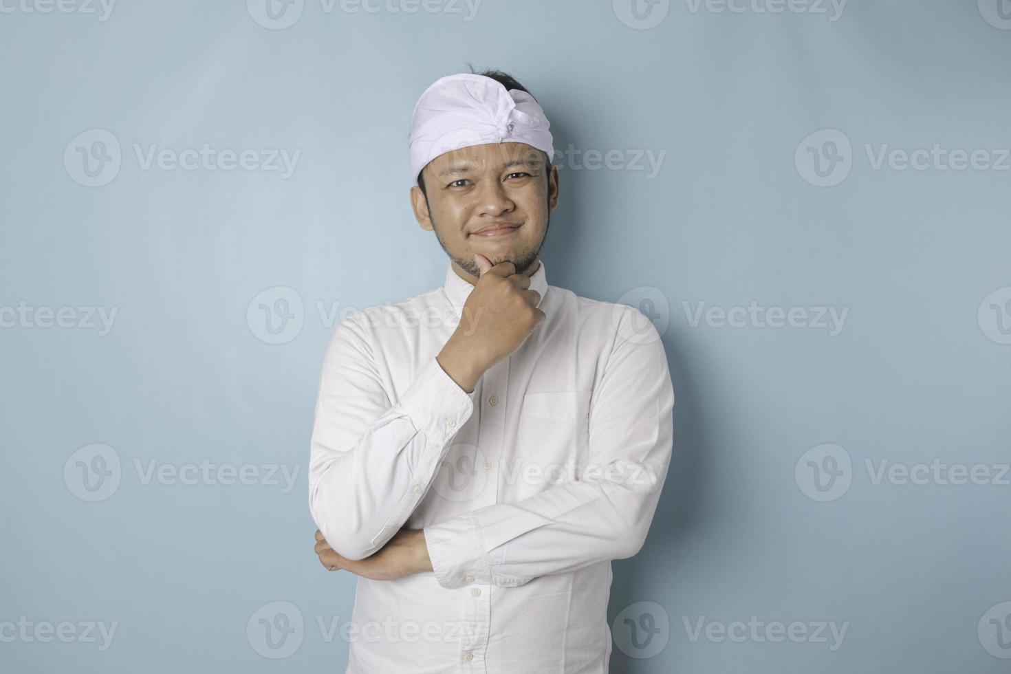 retrato de un joven balinés pensativo que usa udeng o diadema tradicional y camisa blanca mirando a un lado mientras su dedo en la barbilla está aislado sobre fondo azul foto