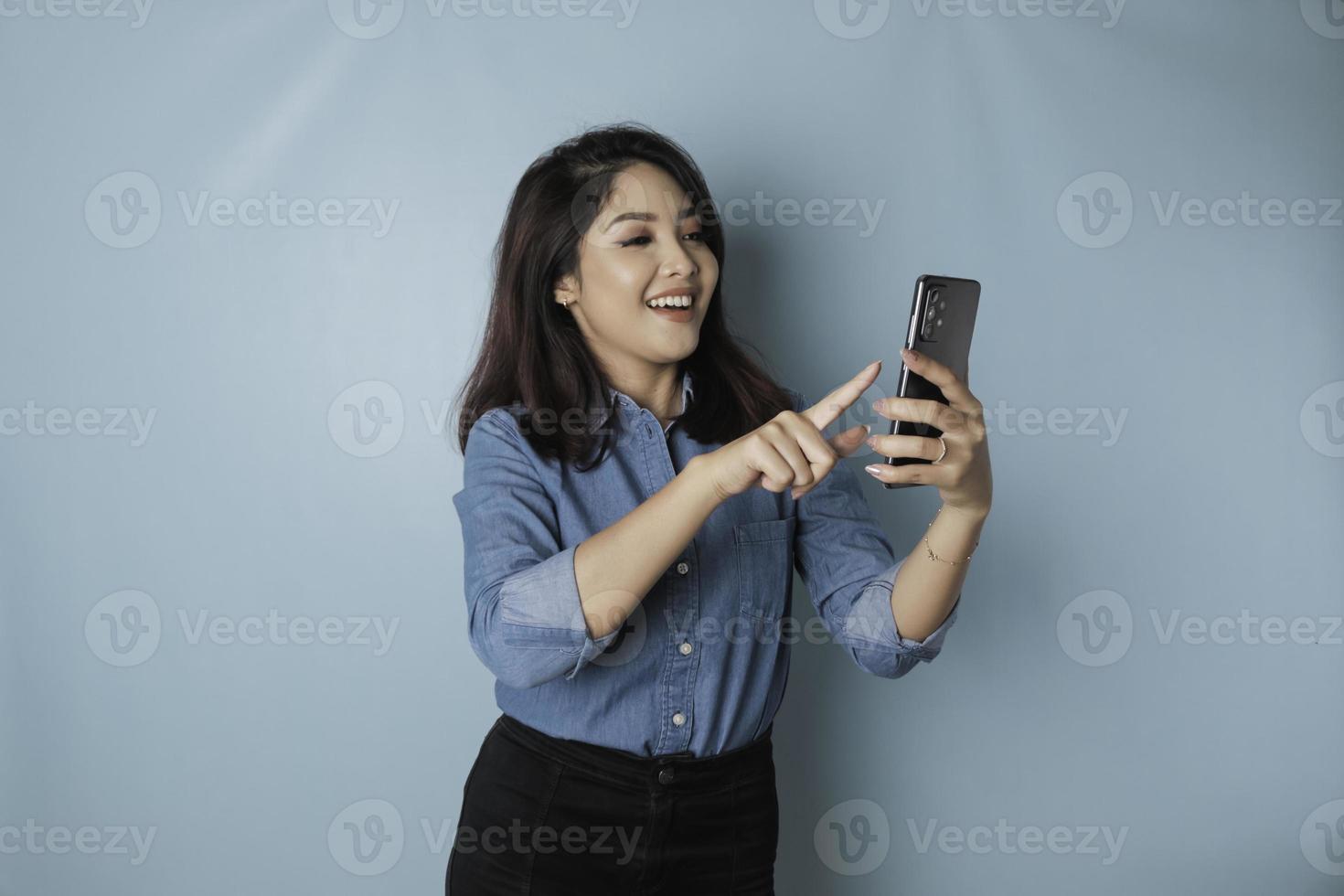 A portrait of a happy Asian woman is smiling and holding her smartphone wearing a blue shirt isolated by a blue background photo