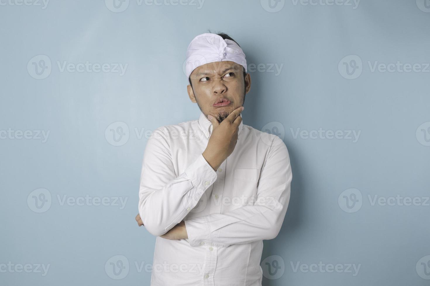 retrato de un joven balinés pensativo que usa udeng o diadema tradicional y camisa blanca mirando a un lado mientras su dedo en la barbilla está aislado sobre fondo azul foto