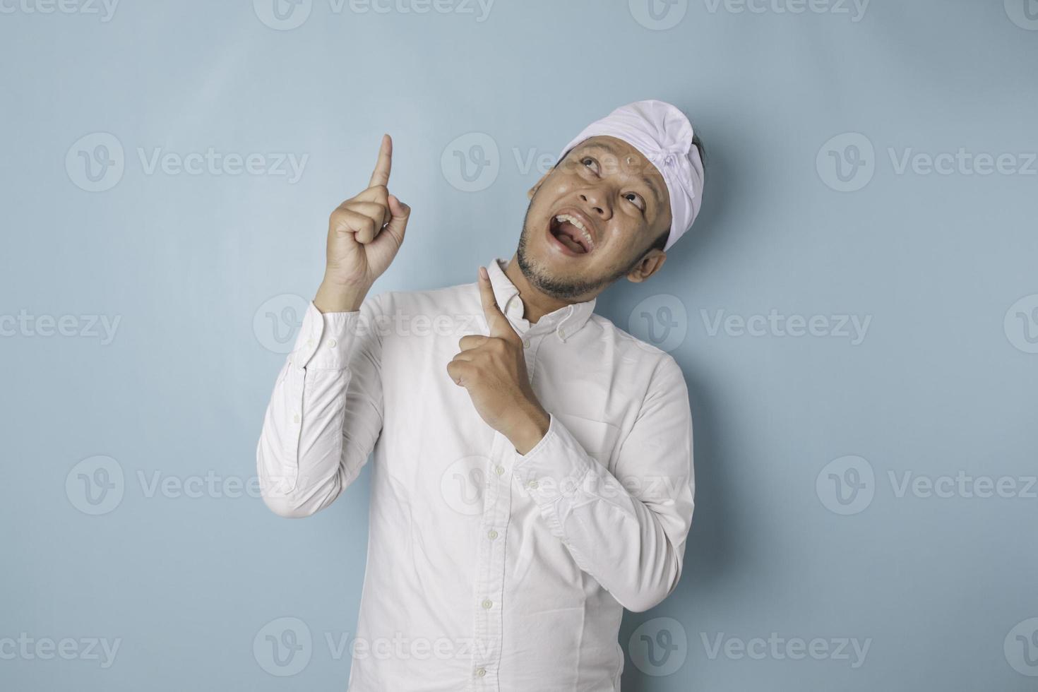 Excited Balinese man wearing udeng or traditional headband and white shirt pointing at the copy space upside him, isolated by blue background photo