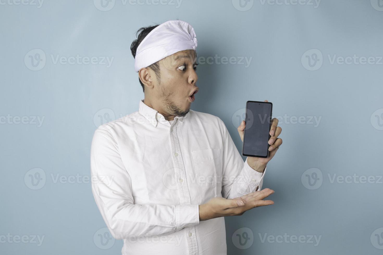 Surprised Balinese man wearing udeng or traditional headband and white shirt holding his smartphone, isolated by blue background photo