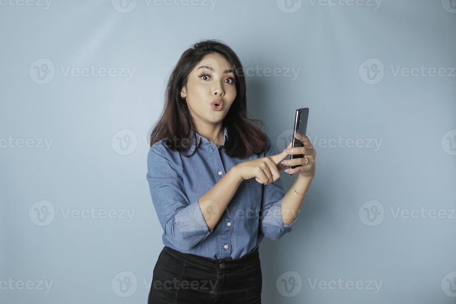 sorprendida mujer asiática con pantalones azul apuntando a su smartphone, aislada de fondo azul foto