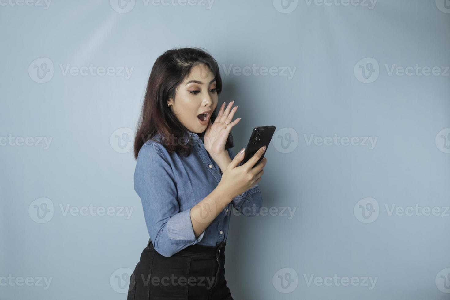 sorprendida mujer asiática con pantalones azul apuntando a su smartphone, aislada de fondo azul foto