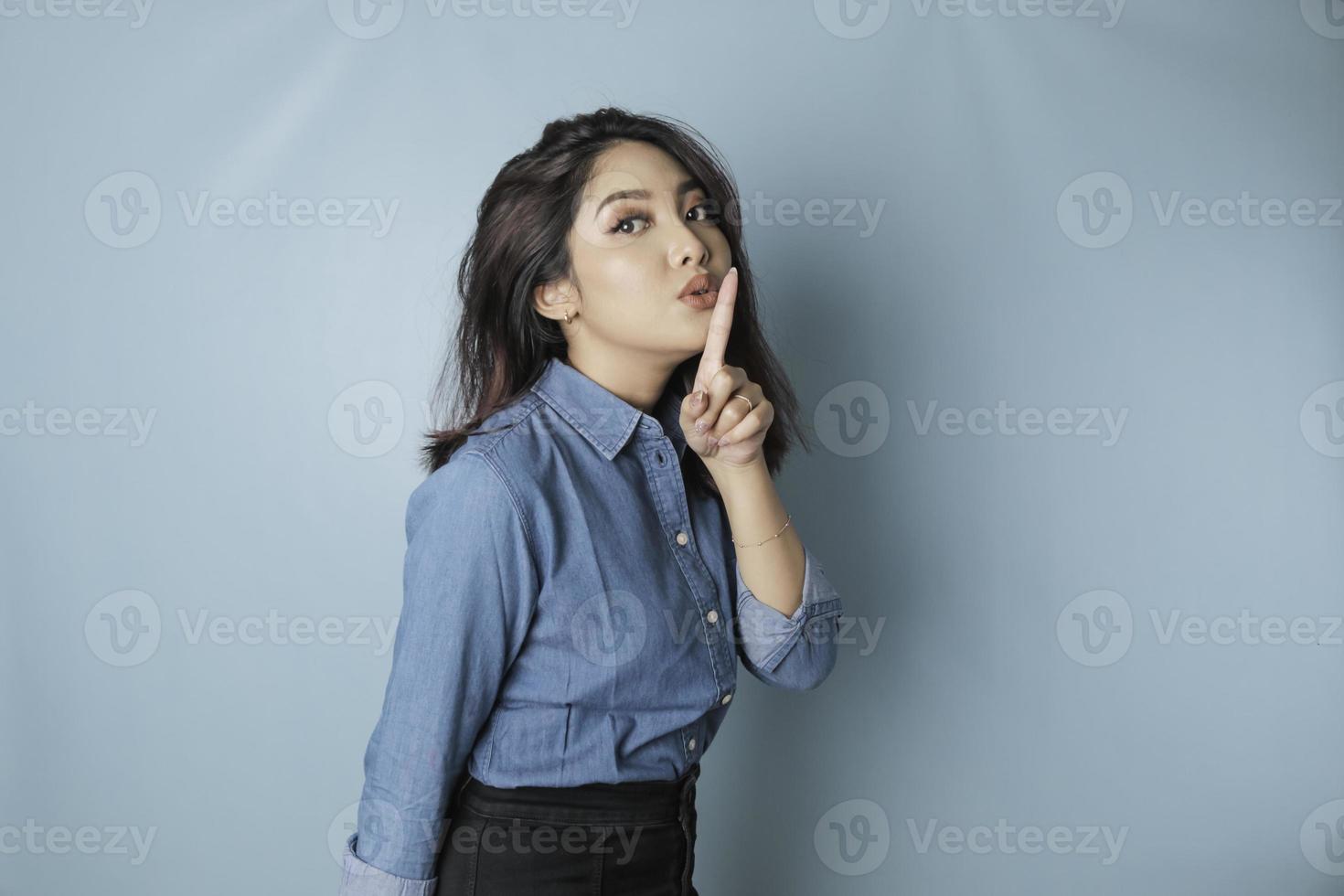 Portrait of a young Asian woman tell be quiet, shushing with serious face, hush with finger pressed to lips, isolated by blue background photo