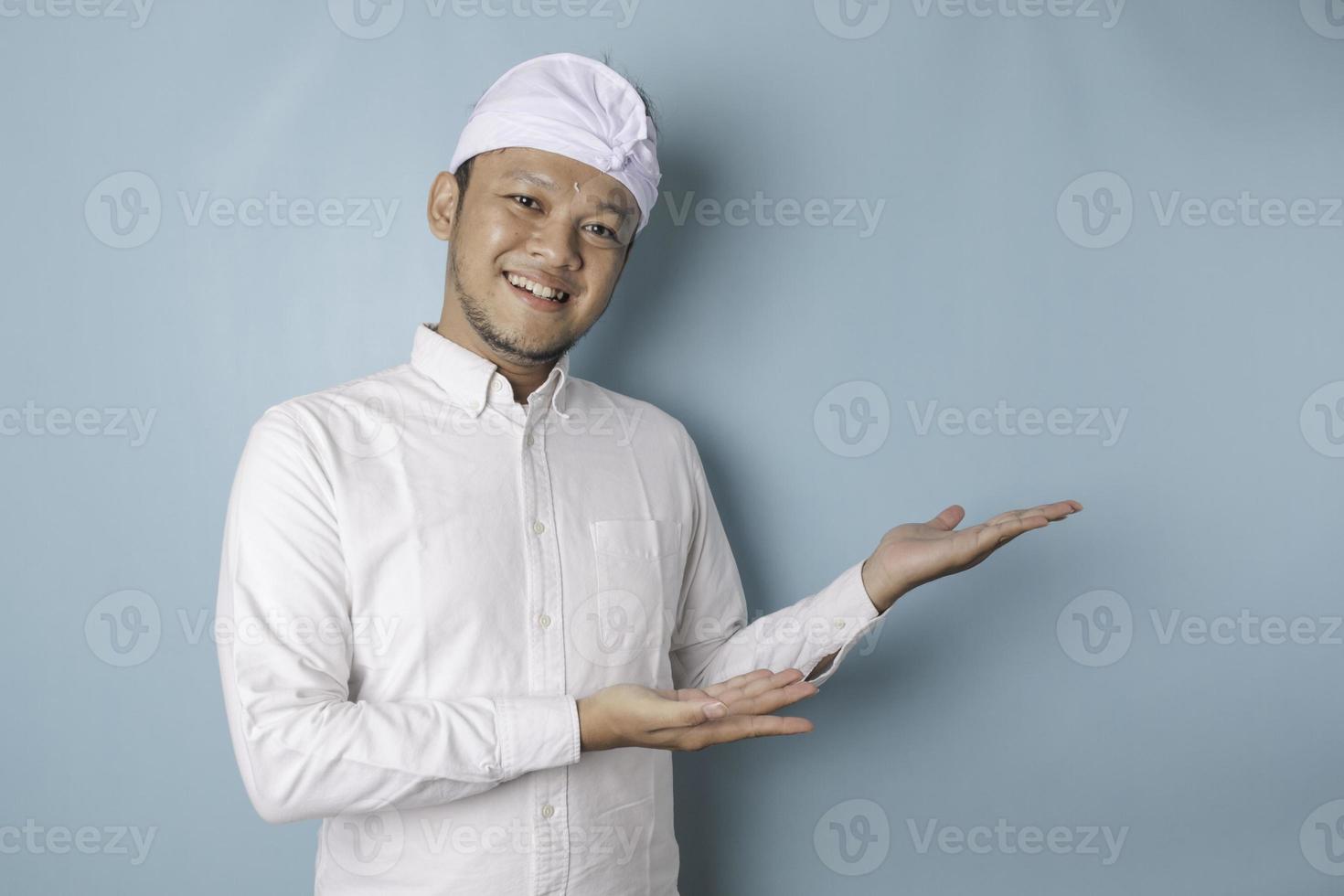 Excited Balinese man wearing udeng or traditional headband and white shirt pointing at the copy space beside him, isolated by blue background photo
