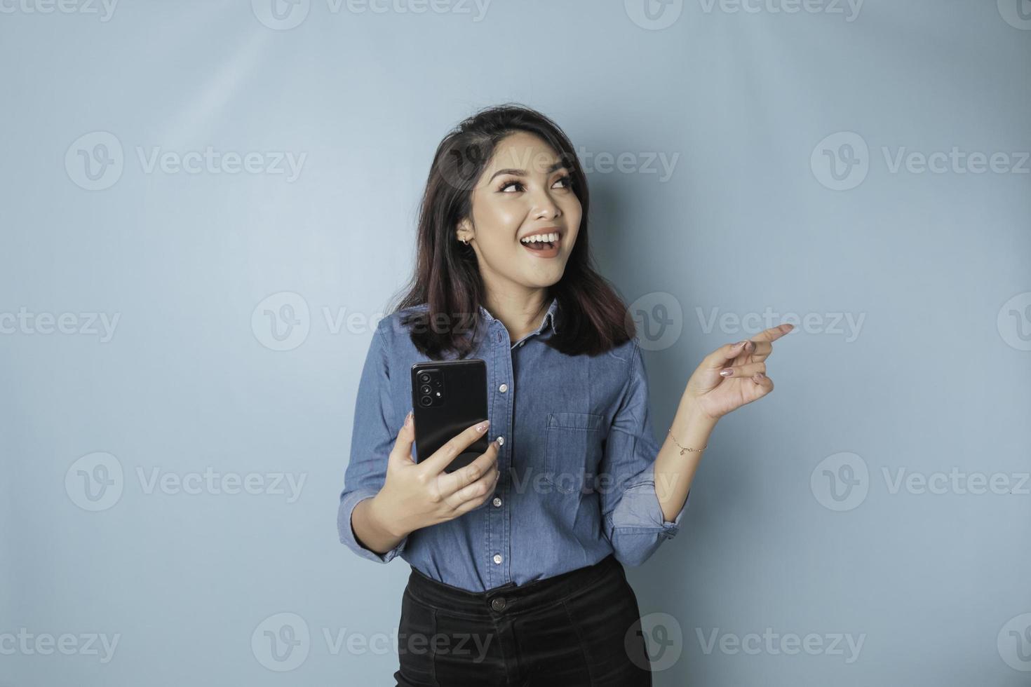 mujer asiática emocionada con pantalones azul apuntando al espacio de copia a su lado mientras sostiene un teléfono inteligente, aislada por fondo azul foto