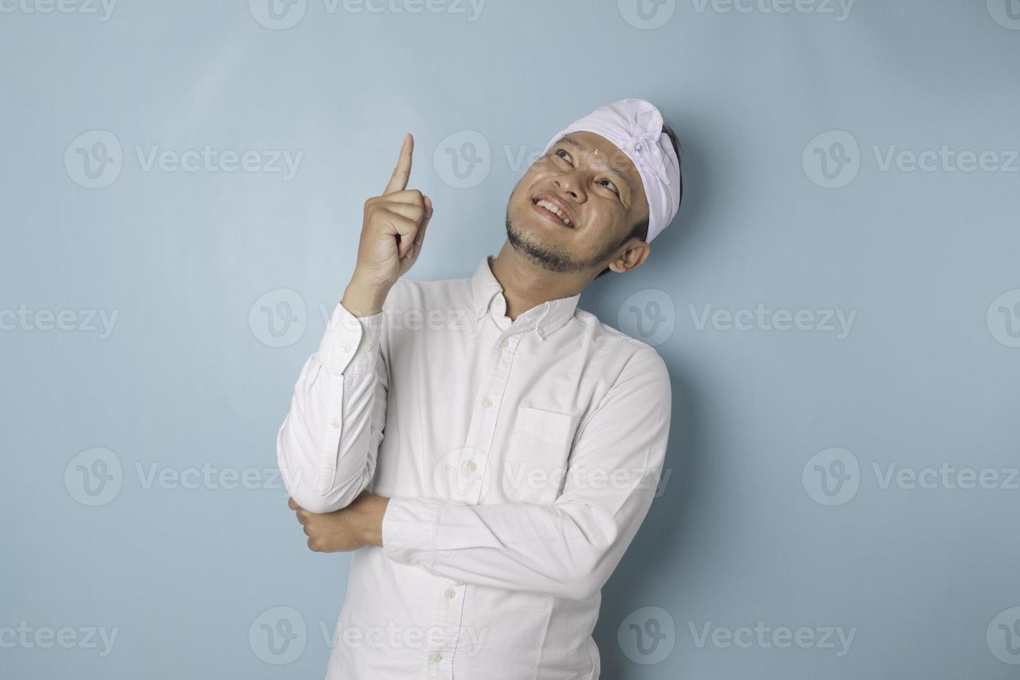 emocionado hombre balinés con udeng o diadema tradicional y camisa blanca apuntando al espacio de la copia al revés, aislado por fondo azul foto