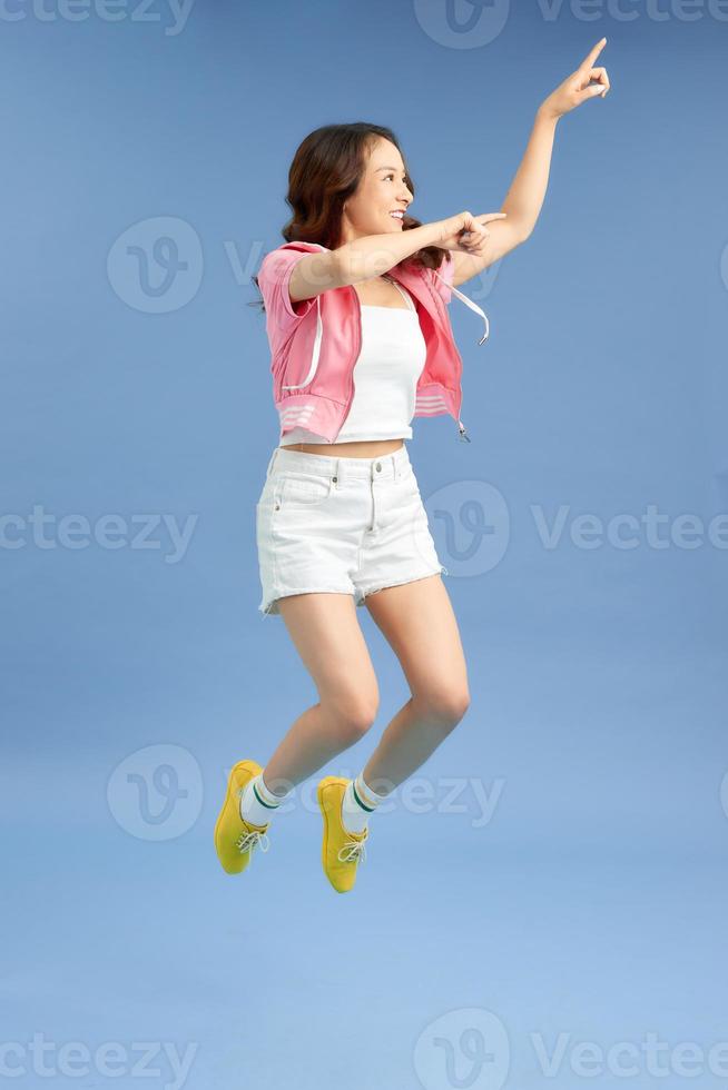 Photo of funny Asian joyful lady in white t-shirt and short jeans jumping and smiling isolated over blue background.