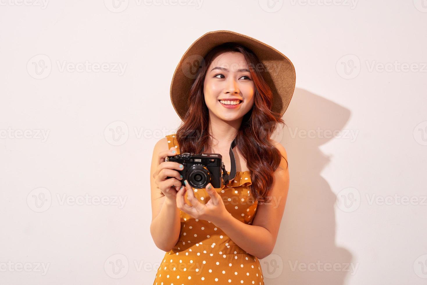 Portrait of cheerful smiling young woman taking photo with inspiration and wearing summer dress. Girl holding retro camera. Model posing on beige background in hat