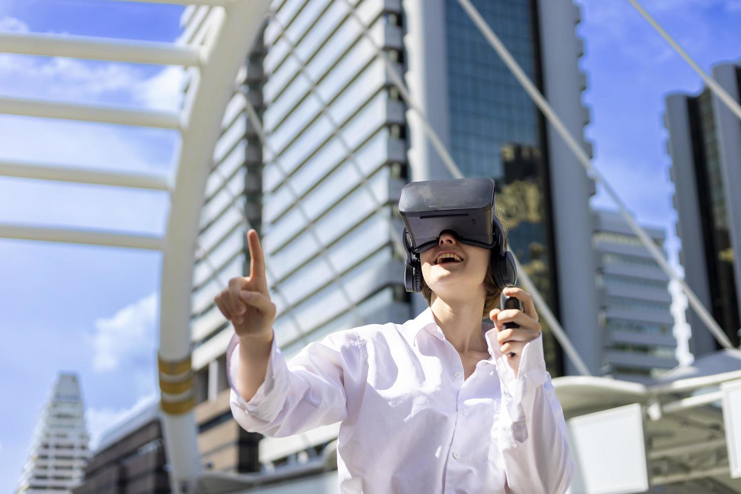 Young caucasian woman wearing VR goggles to play virtual reality simulator game surrounded by modern urban downtown city building photo