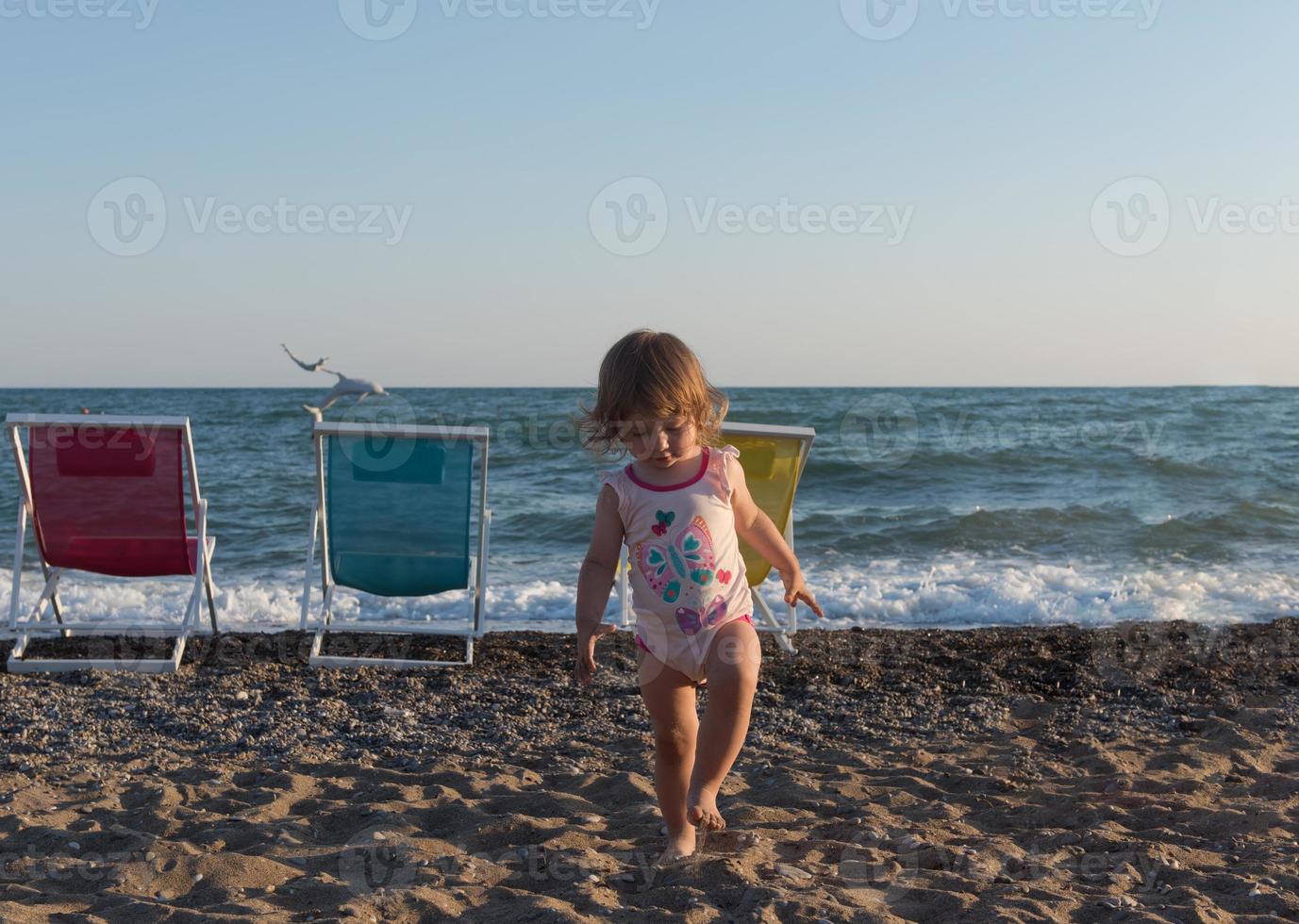 Little girl in sitting on sand and playing with bucket and spade at sea beach in warm sunny summer day. Closeup. Side view. photo