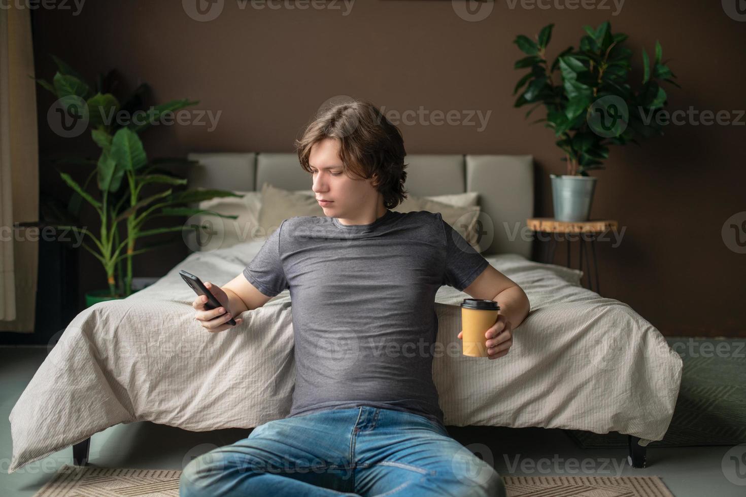 A positive teenage guy sits on the floor of the house in the bedroom with a cup of coffee in an eco-craft glass, relaxes and communicates on the phone. photo