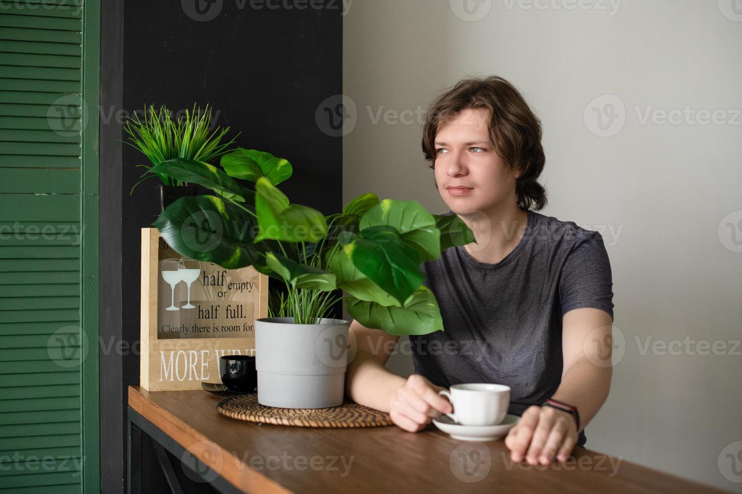 A positive teenage guy sits at the table at home in the kitchen with a cup of coffee in a glass, relaxes and rests in the morning. photo
