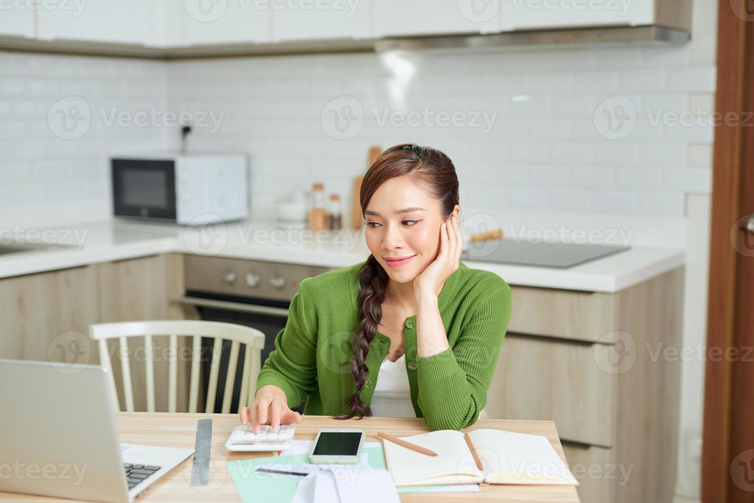 Attractive young smiling Woman paying bills online using laptop from home while sitting in the kitchen photo