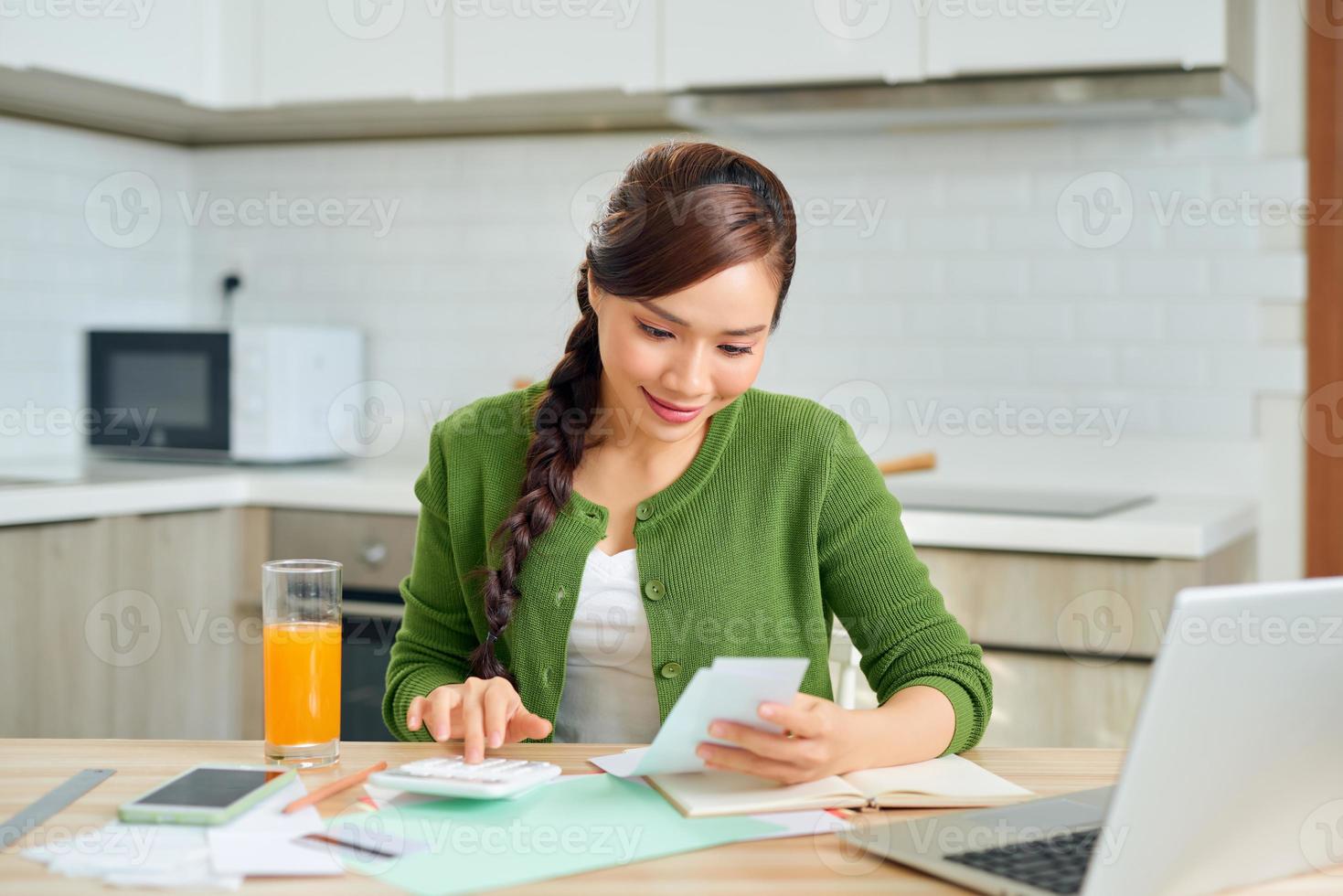 atractiva joven sonriente pagando facturas en línea usando una laptop desde casa mientras se sienta en la cocina foto