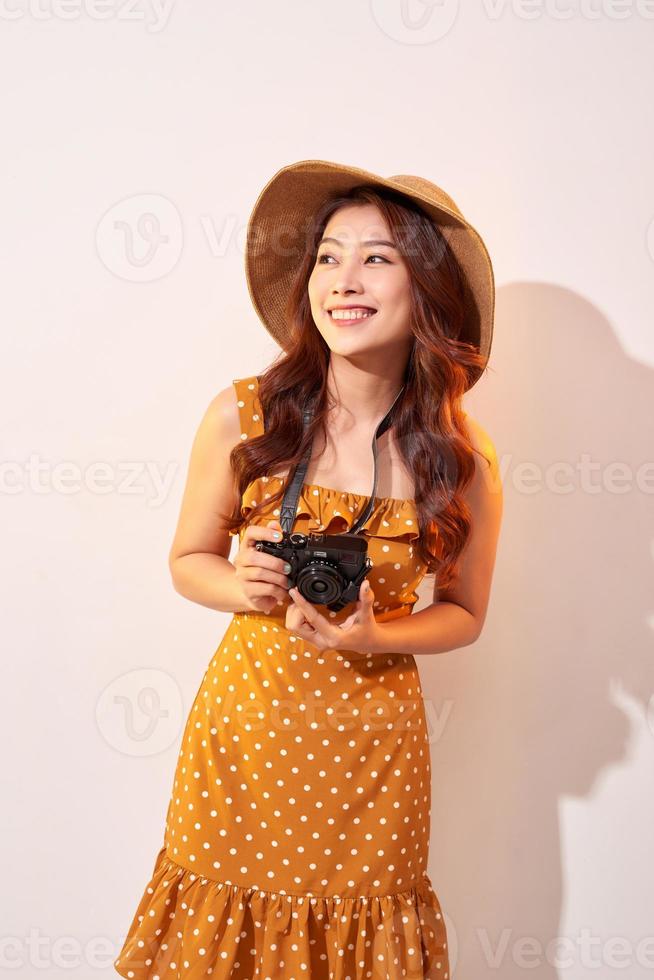 Portrait of cheerful smiling young woman taking photo with inspiration and wearing summer dress. Girl holding retro camera. Model posing on beige background in hat