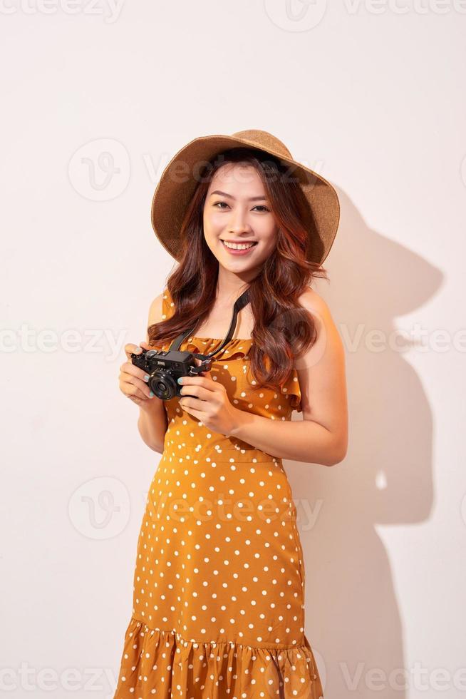 Portrait of cheerful smiling young woman taking photo with inspiration and wearing summer dress. Girl holding retro camera. Model posing on beige background in hat