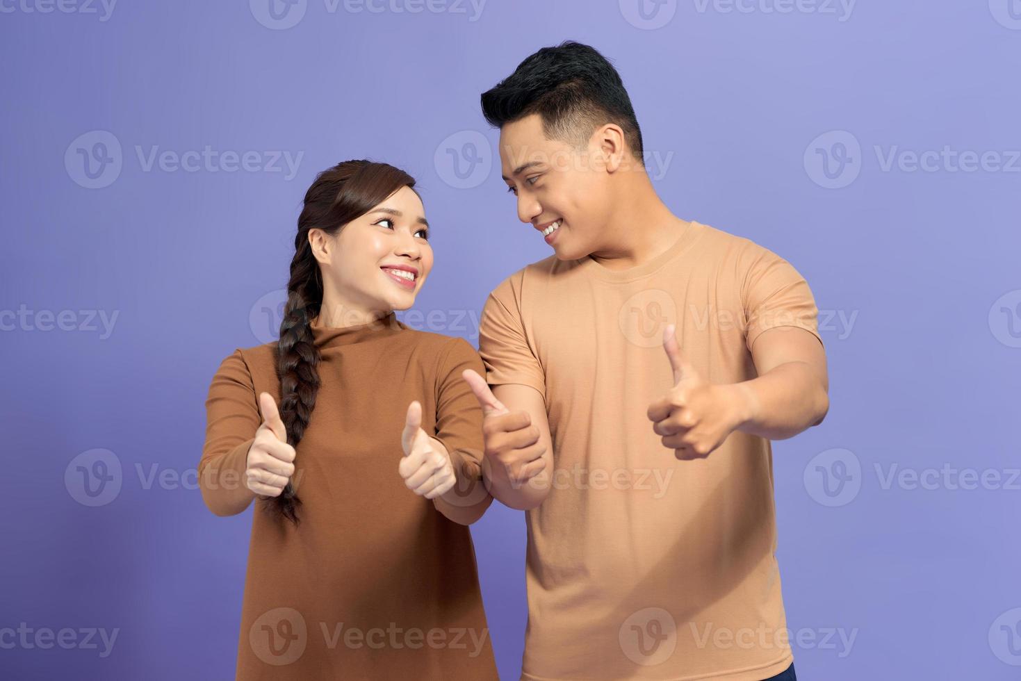 feliz pareja joven y encantadora mostrando los pulgares hacia arriba y mirando a la cámara sobre fondo púrpura foto