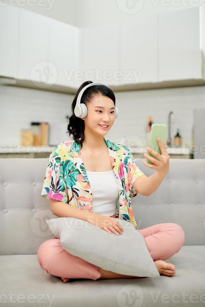 Portrait beautiful young asian woman use smart mobile phone with headphone for listen music on sofa in living room area photo