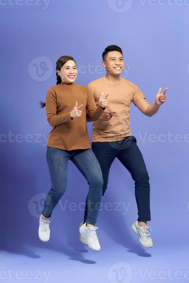 Energetic beautiful couple posing together on camera while running or jumping and pointing fingers on copyspace along background photo