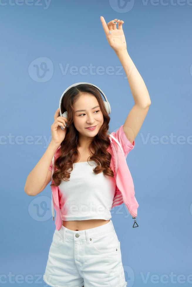 Chica asiática de energía con auriculares escuchando música con los ojos cerrados sobre fondo azul en el estudio. ella usa camiseta blanca, pantalones cortos. foto