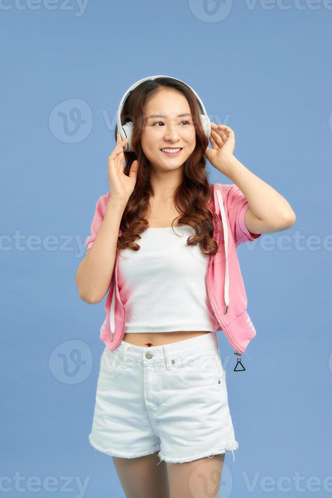 Chica asiática de energía con auriculares escuchando música con los ojos cerrados sobre fondo azul en el estudio. ella usa camiseta blanca, pantalones cortos. foto