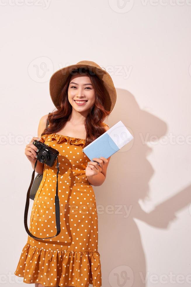 Portrait of a happy young woman in hat holding camera and showing passport while standing isolated over beige background photo