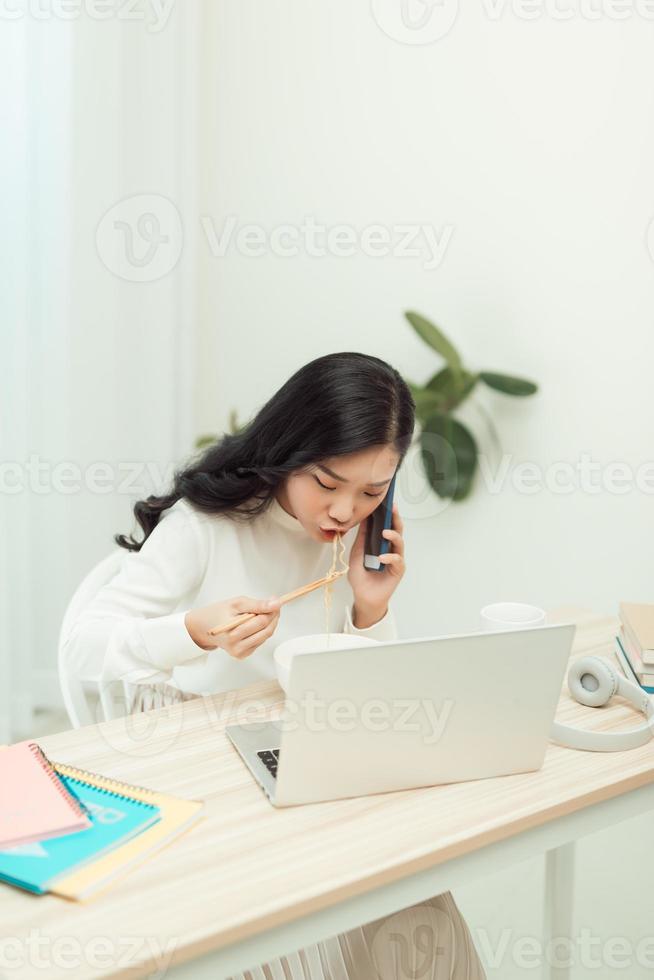 Confident smiling attractive young asian woman entrepreneur having lunch break while sitting at the office desk and using mobile phone photo