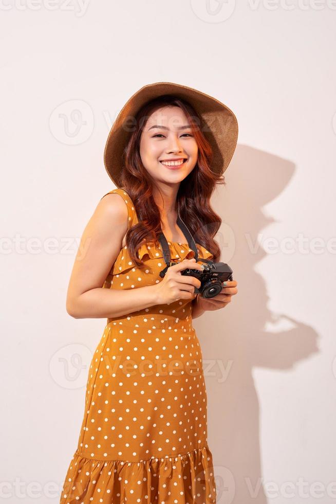 Portrait of cheerful smiling young woman taking photo with inspiration and wearing summer dress. Girl holding retro camera. Model posing on beige background in hat