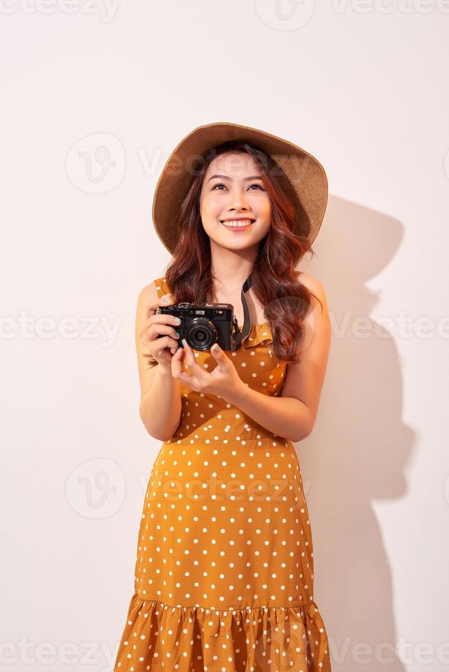 Portrait of cheerful smiling young woman taking photo with inspiration and wearing summer dress. Girl holding retro camera. Model posing on beige background in hat