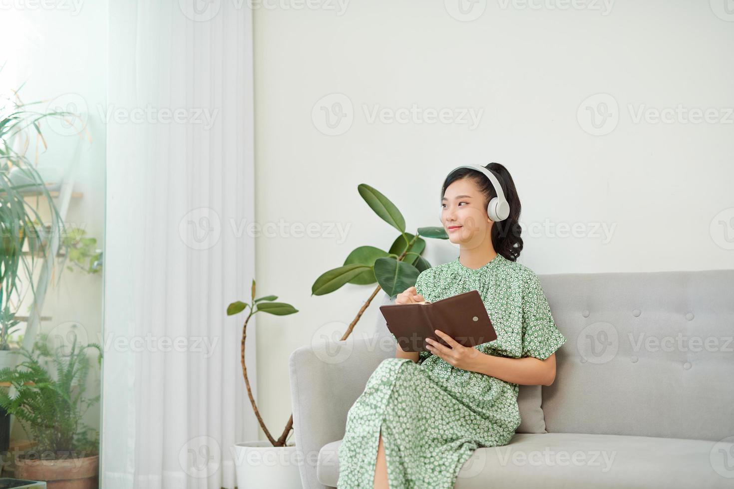 mage of focused asian woman using headphones and writing down notes while sitting on sofa at home photo