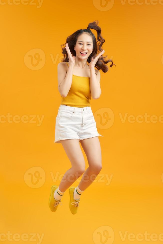 Joyful young asian girl having fun fooling around jumping doing winner gesture on yellow background. photo