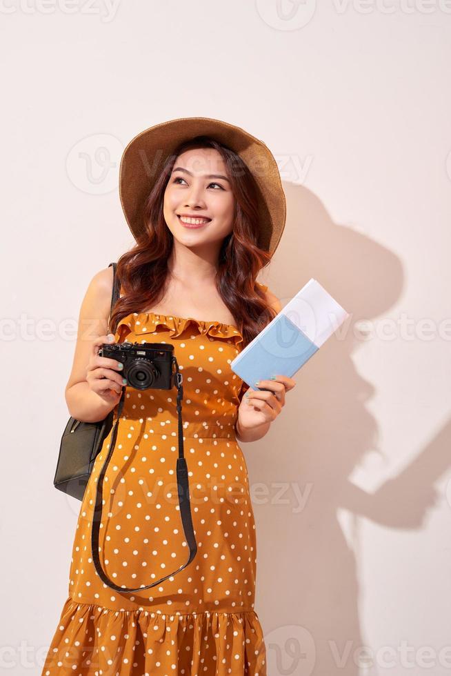 Portrait of a happy young woman in hat holding camera and showing passport while standing isolated over beige background photo