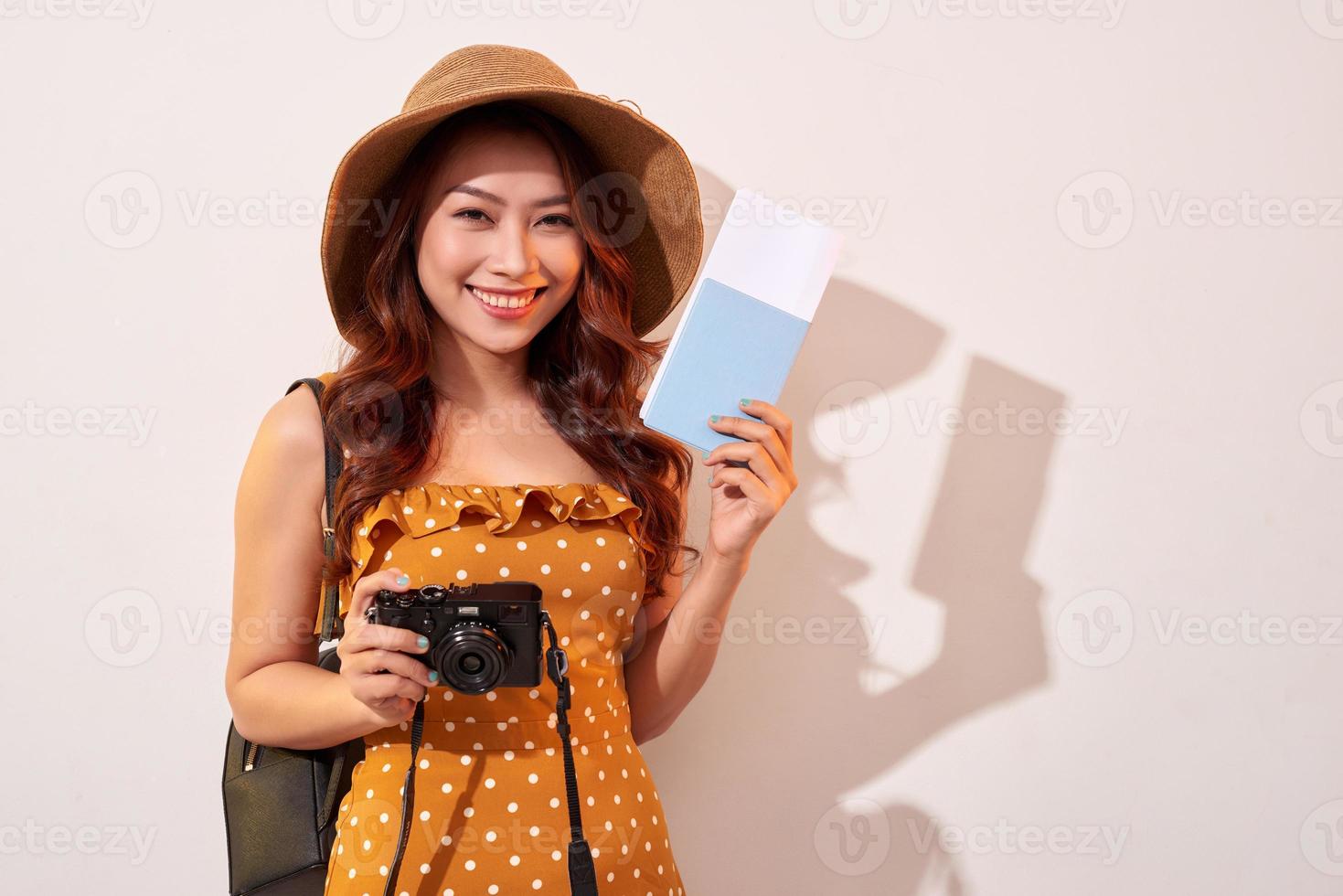 Portrait of a happy young woman in hat holding camera and showing passport while standing isolated over beige background photo