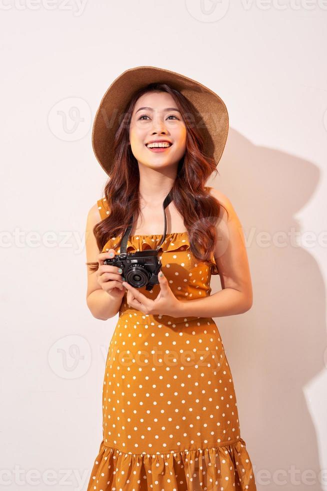 Portrait of cheerful smiling young woman taking photo with inspiration and wearing summer dress. Girl holding retro camera. Model posing on beige background in hat
